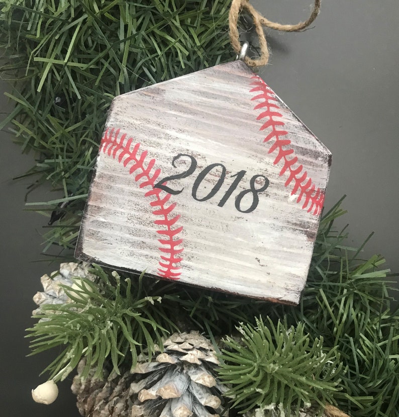 Pu&ograve; includere: Un ornamento di Natale in legno bianco a forma di baseball con cuciture rosse e l'anno 2018 dipinto su di esso.