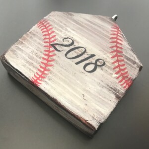 Pu&ograve; includere: Un ornamento a forma di home plate di baseball in legno bianco con cuciture rosse e l'anno 2018 dipinto su di esso.