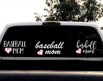 May include: A white truck with a black rear window. The window has three decals that say "baseball mom" with a red heart and a baseball inside the heart.