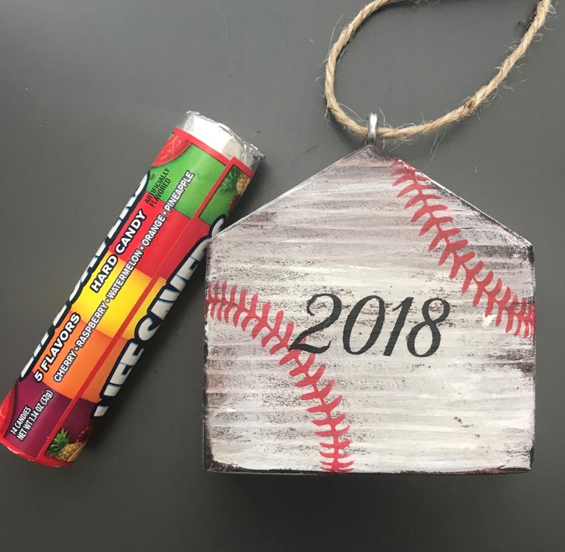 Pu&ograve; includere: Un ornamento per la casa in legno bianco a forma di piatto di casa da baseball con cuciture rosse e l'anno 2018 dipinto su di esso. L'ornamento ha un gancio in corda.