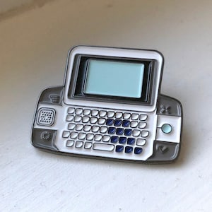 Może przedstawiać: Szpilka emaliowana w kolorze szarym i białym przedstawiająca zabytkowy telefon komórkowy z małym ekranem i klawiaturą. Szpilka ma kolorystykę niebiesko-białą.