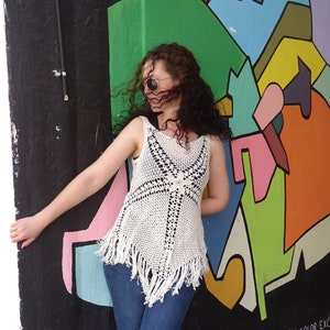 May include: A woman wearing a white crocheted top with fringe, blue jeans, and sunglasses, standing in front of a colorful wall with abstract shapes and the text "#THE COLOR EXCHANGE".