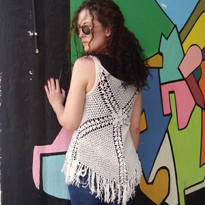 May include: A white crochet top with a fringe hem. The top is sleeveless and has a loose fit. The model is wearing blue jeans.