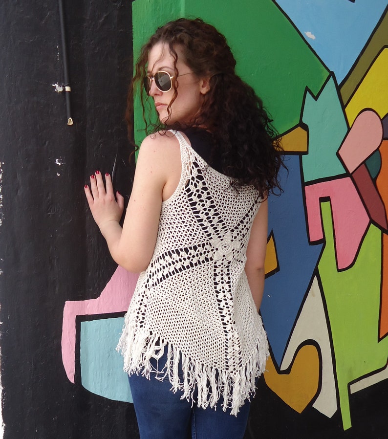 May include: A white crochet top with a fringe hem. The top is sleeveless and has a loose fit. The model is wearing blue jeans.