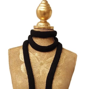 May include: A black knitted scarf displayed on a gold mannequin. The scarf is long and narrow, with a simple, ribbed texture. The mannequin has a gold finish and a decorative top.