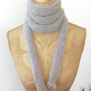 May include: A pale grey knitted scarf displayed on a mannequin. The scarf is made from a soft, fluffy yarn and has a simple, rectangular design. It is wrapped around the mannequin's neck, with two long sections hanging down.