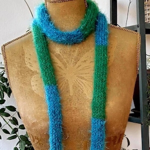 Op de afbeelding: Een zachte, handgemaakte sjaal in blauw- en groentinten. De sjaal wordt getoond op een mannequin, die de zachte textuur en het colorblock-ontwerp laat zien. Een gezellig accessoire.