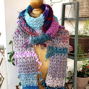 Puede incluir: Bufanda de ganchillo multicolor, expuesta sobre un maniquí. La bufanda presenta hilos azules, morados y grises con flecos y detalles texturizados. Un accesorio acogedor para el clima fresco, hecho a mano.
