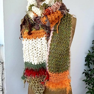 Puede incluir: Bufanda hecha a mano, colorida, exhibida en un maniquí. La bufanda presenta una variedad de texturas y colores, incluyendo blanco, verde, naranja y rojo. El diseño incorpora elementos tejidos y de ganchillo, con secciones con flecos y texturizadas.