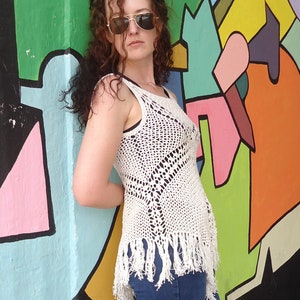 May include: A woman wearing a white crocheted top with fringe detailing and blue jeans. She is standing in front of a colorful wall with abstract shapes.
