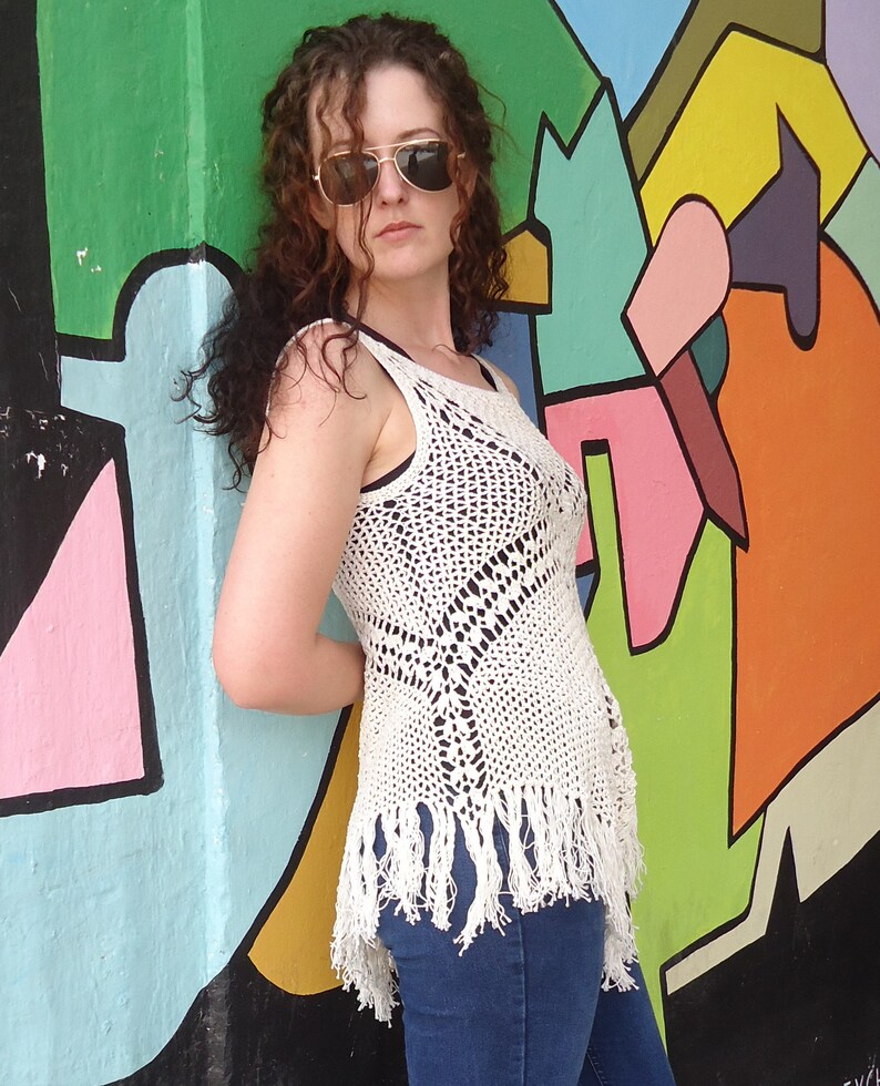 May include: A woman wearing a white crocheted top with fringe detailing and blue jeans. She is standing in front of a colorful wall with abstract shapes.
