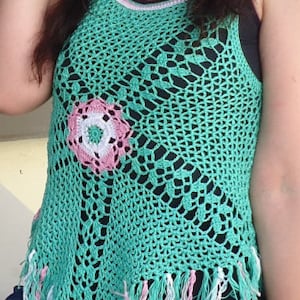 May include: A green crochet top with a pink and white flower design in the center. The top has a fringe trim at the bottom.