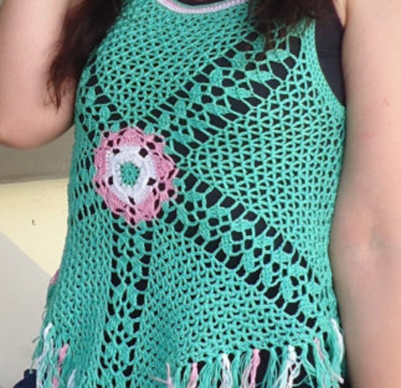 May include: A green crochet top with a pink and white flower design in the center. The top has a fringe trim at the bottom.
