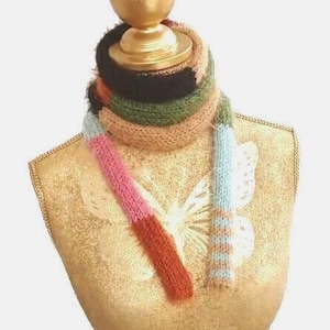 Könnte beinhalten: Ein mehrfarbiger Strickschal mit horizontalen Streifen in Schwarz, Olivgrün, Hellbraun, Rosa, Hellblau und Rostorange. Der Schal ist um den Hals eines goldenen Schneiderbüsten-Torsos gewickelt, wobei zwei lange Enden vorne am Körper herabhängen.