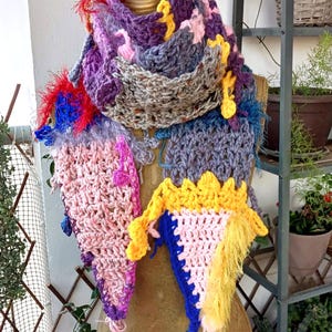 Può includere: Una sciarpa colorata all'uncinetto fatta a mano, esposta su un manichino. La sciarpa presenta una varietà di colori tra cui viola, rosa, giallo e blu, con motivi strutturati e dettagli con frange. Un accessorio artistico e unico.