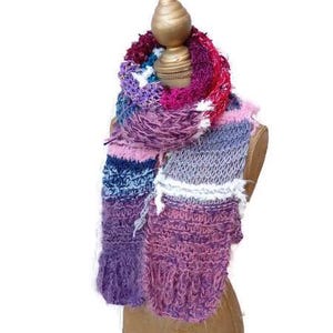 Könnte beinhalten: Ein mehrfarbiger gestrickter Schal mit einer Mischung aus Texturen, darunter flauschiges und dickes Garn. Der Schal ist in den Farben Rosa, Lila, Blau und Weiß.