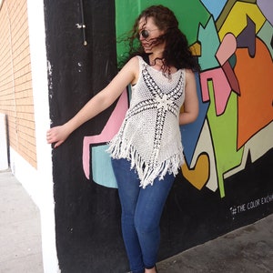 May include: A woman wearing a white crocheted top with fringe, blue jeans, and sunglasses stands in front of a colorful wall with abstract shapes and the text "#THE COLOR EXCHANGE".