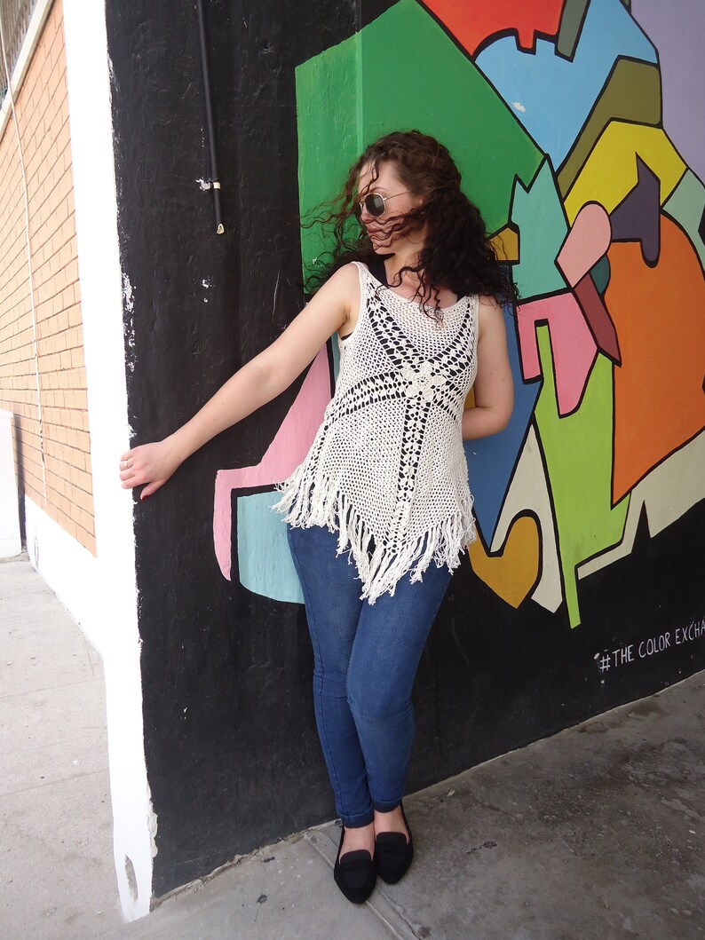 May include: A woman wearing a white crocheted top with fringe, blue jeans, and sunglasses stands in front of a colorful wall with abstract shapes and the text "#THE COLOR EXCHANGE".
