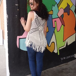 May include: A woman wearing a white crocheted top with fringe, blue jeans, and sunglasses, standing in front of a colorful wall with the text "#THE COLOR EXCHANGE".