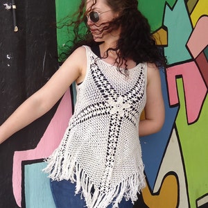 May include: A white crochet top with fringe detailing. The top is sleeveless and has a loose fit. The model is wearing the top with blue jeans.