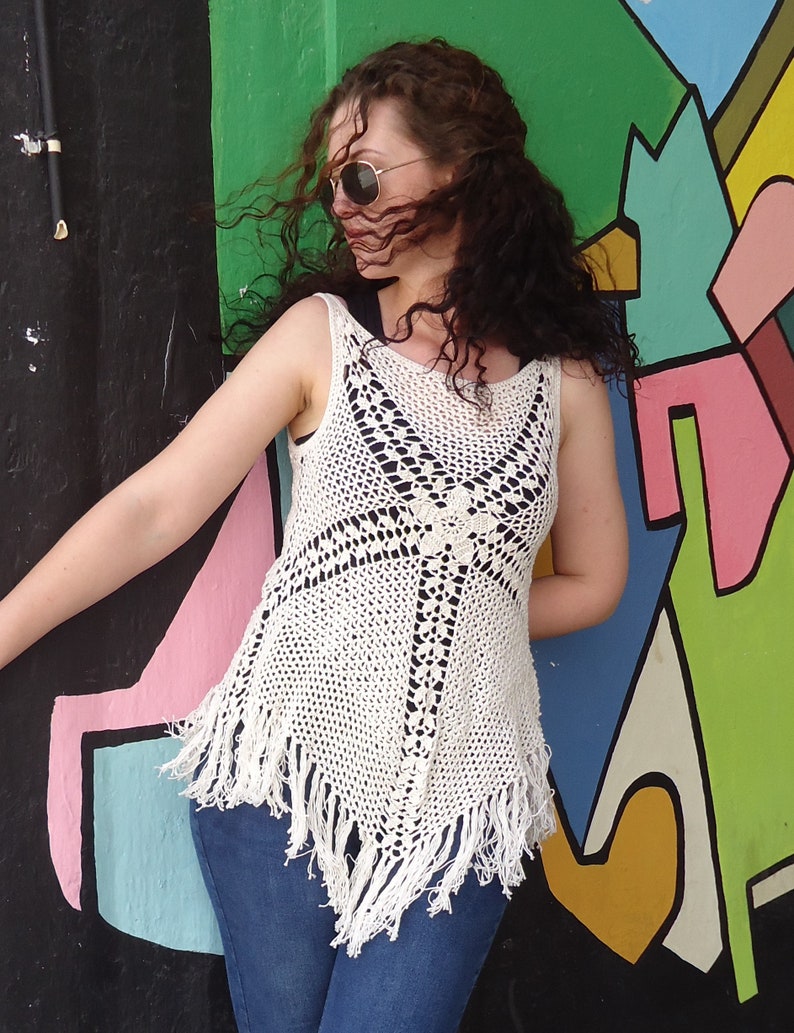 May include: A white crochet top with fringe detailing. The top is sleeveless and has a loose fit. The model is wearing the top with blue jeans.