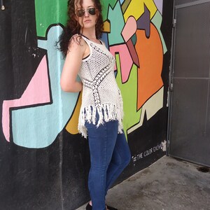 May include: A woman wearing a white crocheted top with fringe, blue jeans, and sunglasses. She is standing in front of a colorful wall with the text "#THE COLOR EXCHANGE" on it.