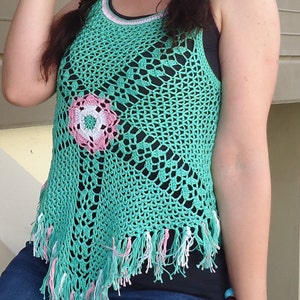 May include: A green crochet top with a pink and white flower design. The top has a fringed hem and is sleeveless.