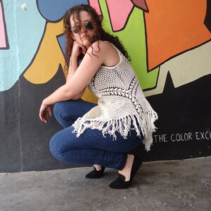 May include: A woman wearing a white crocheted top with fringe, blue jeans, and black shoes. She is posing in front of a colorful wall with the text "THE COLOR EXC" visible.