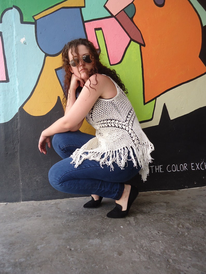 May include: A woman wearing a white crocheted top with fringe, blue jeans, and black shoes. She is posing in front of a colorful wall with the text "THE COLOR EXC" visible.
