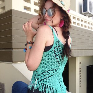 May include: A woman wearing a turquoise crochet top with fringe, a straw hat with pink flowers, and blue jeans. She is wearing sunglasses and pink earrings.