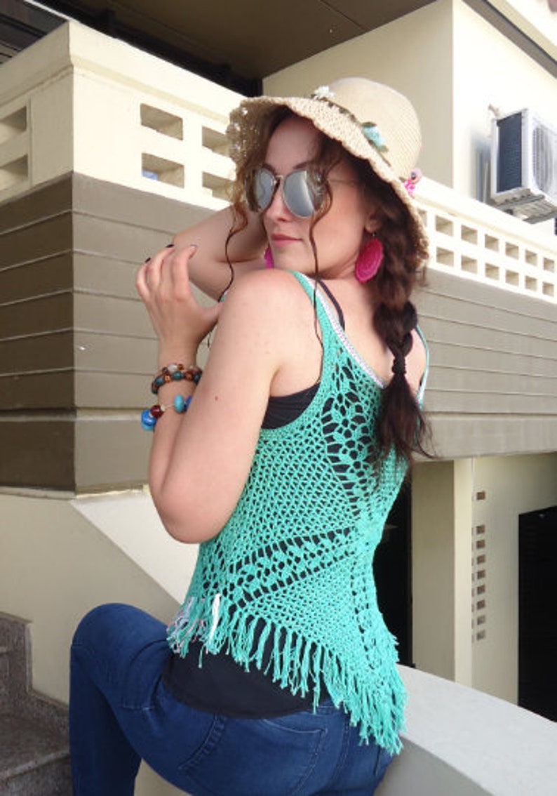 May include: A woman wearing a turquoise crochet top with fringe, a straw hat with pink flowers, and blue jeans. She is wearing sunglasses and pink earrings.