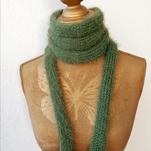 May include: A green knitted scarf is draped over a mannequin. The scarf is a soft, fuzzy material and has a long, rectangular shape. The mannequin is a light brown color and has a butterfly design on the front.