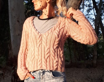 May include: A light pink cable-knit sweater with a deep V-neck. The sweater has long sleeves and a cropped fit. The model is wearing acid wash jeans and sunglasses. The background features trees and natural light.