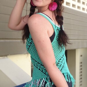 May include: A woman wearing a turquoise crochet top with fringe, a straw hat, and blue jeans. She is wearing sunglasses and pink earrings.