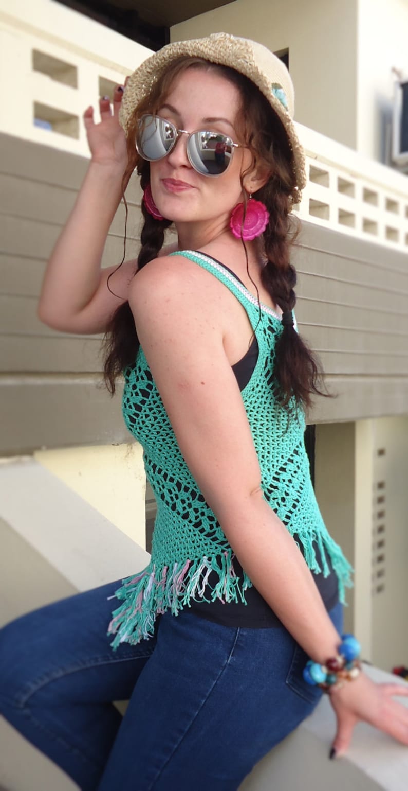 May include: A woman wearing a turquoise crochet top with fringe, a straw hat, and blue jeans. She is wearing sunglasses and pink earrings.