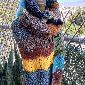 May include: A multi-coloured crochet scarf featuring a patchwork design with varied yarn textures and colours including blue, yellow, brown, green, and black. The scarf is draped over a mannequin, displaying a thick, chunky knit construction.