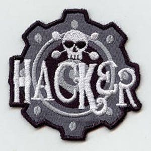 May include: A grey embroidered patch featuring a gear design with a skull and crossbones in the centre. The word "HACKER" is written in white around the skull.