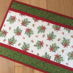 May include: A rectangular Christmas table runner with a white center featuring a pattern of green sprigs, red berries, and bows. The edges are bordered with red and green patterned fabric. The runner is quilted.