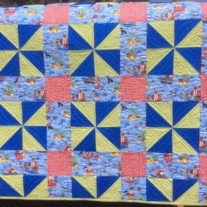 May include: A handmade quilt with a nautical theme. The quilt features squares of blue fabric with pirate ship and island scenes, alternating with red gingham and pinwheel designs in blue and green. The quilt is likely made of cotton.