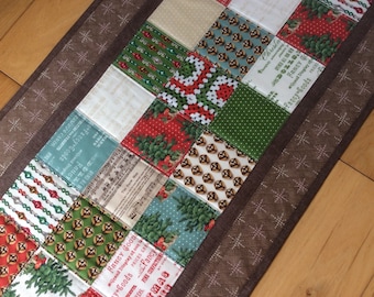 May include: A festive Christmas table runner with a patchwork design. The runner features a brown border and squares with various patterns, including Christmas trees and text. The table runner is approximately 30 cm wide, perfect for the holiday season.