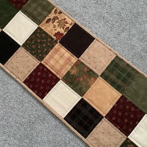 Puede incluir: Camino de mesa acolchado con diseño de patchwork. El camino de mesa presenta cuadrados en varios colores, incluyendo beige, verde, burdeos y negro. Algunos cuadrados tienen patrones de hojas. Los bordes están ribeteados con una tela marrón claro.