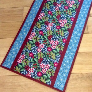 May include: A rectangular table runner with a floral pattern. The center features a dark blue background with red flowers, green leaves, and red berries. The edges are bordered by a burgundy fabric and light blue fabric with a small floral design.