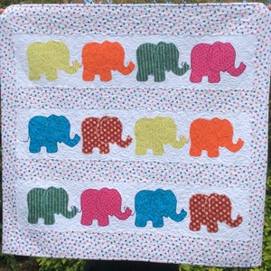 Puede incluir: Un edredón colorido con filas de elefantes aplicados en varios patrones y colores, incluyendo amarillo, naranja, verde, rosa y azul. El edredón tiene un fondo blanco con un borde con estampado de estrellas.