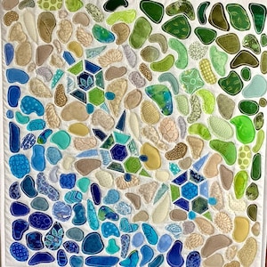 May include: A square textile wall hanging featuring a mosaic of numerous fabric shapes in shades of blue, green, and beige. The composition includes three distinct geometric turtle motifs integrated among the organic, pebble-like fabric pieces.
