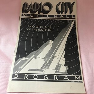 May include: Black and white illustration of Radio City Music Hall, a tall building with a rounded top, with the text "Radio City Music Hall Show Place of the Nation Program Harold K. Simon" at the bottom.