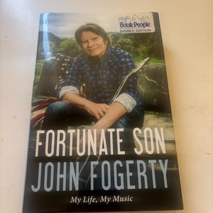 May include: A hardcover book titled Fortunate Son by John Fogerty, My Life, My Music. The cover features a photograph of John Fogerty in a blue plaid shirt holding a guitar, with a Book People Signed Edition sticker in the upper right corner.