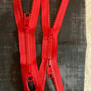 Puede incluir: Dos cremalleras rojas con deslizadores metálicos. Las cremalleras están dispuestas sobre un fondo de tela gris oscuro.