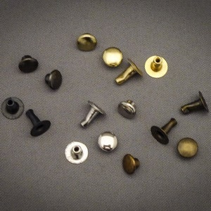 Può includere: Assortimento di rivetti metallici in vari colori, tra cui argento, oro, nero e bronzo. I rivetti sono di diverse dimensioni e forme, alcuni con testa piatta e altri con testa bombata.