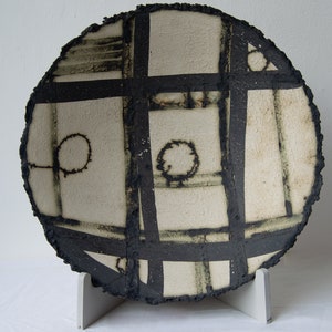 May include: A white ceramic plate with a black geometric design. The plate has a rough, textured surface and is displayed on a white stand.