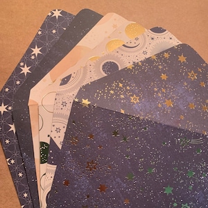 May include: A set of five decorative envelopes with a celestial theme. The envelopes are a dark purple color with gold and green foil accents. The designs feature stars, moons, and other celestial bodies.